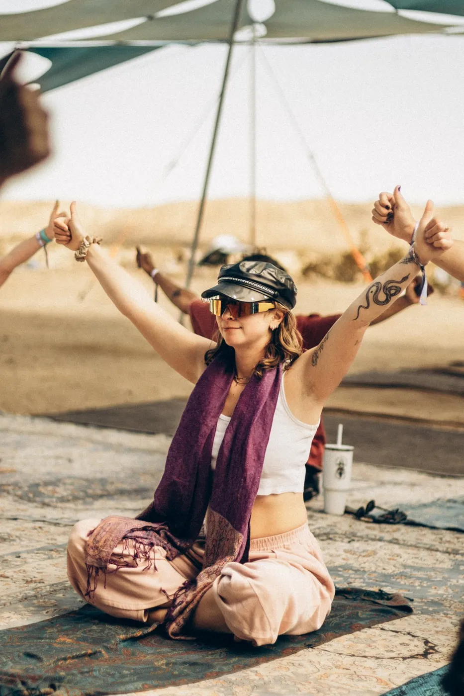 A woman sitting on a mat in the desert with both arms raised, smiling